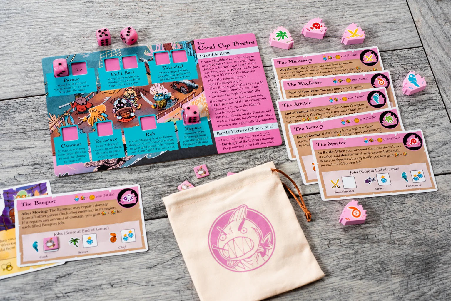 Ahoy: Fang & Fortune board game expansion components for "The Coral Cap Pirates," including action cards, pink dice, and a cloth bag with a shark logo