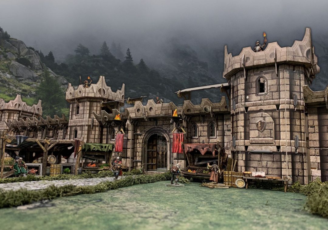 A miniature medieval castle diorama, with guards on towers and armed figures around market stalls, against foggy mountains.