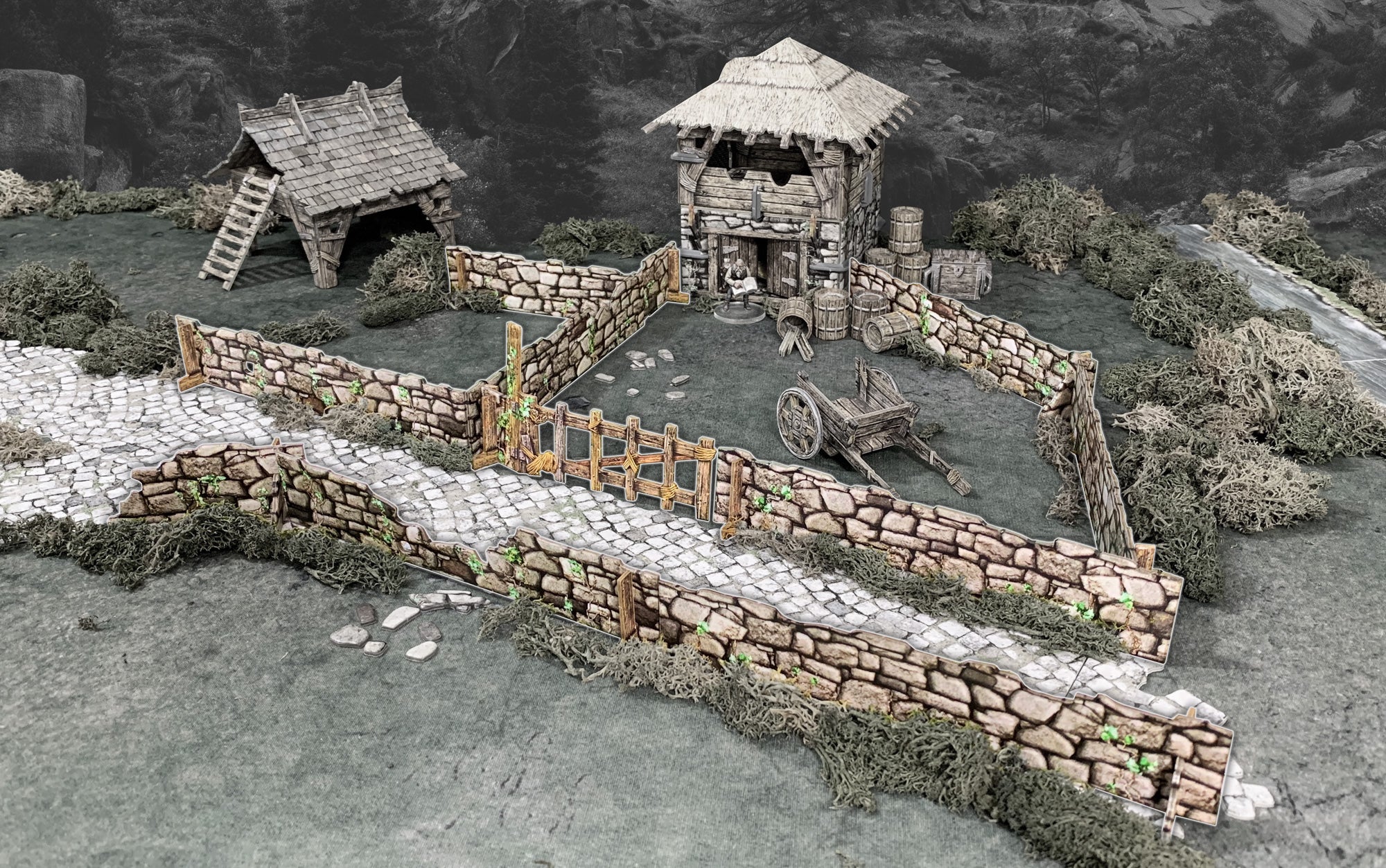 Tabletop terrain of a medieval village: paved road, stone walls, thatched tower, small hut, wooden cart, and a standing figure.
