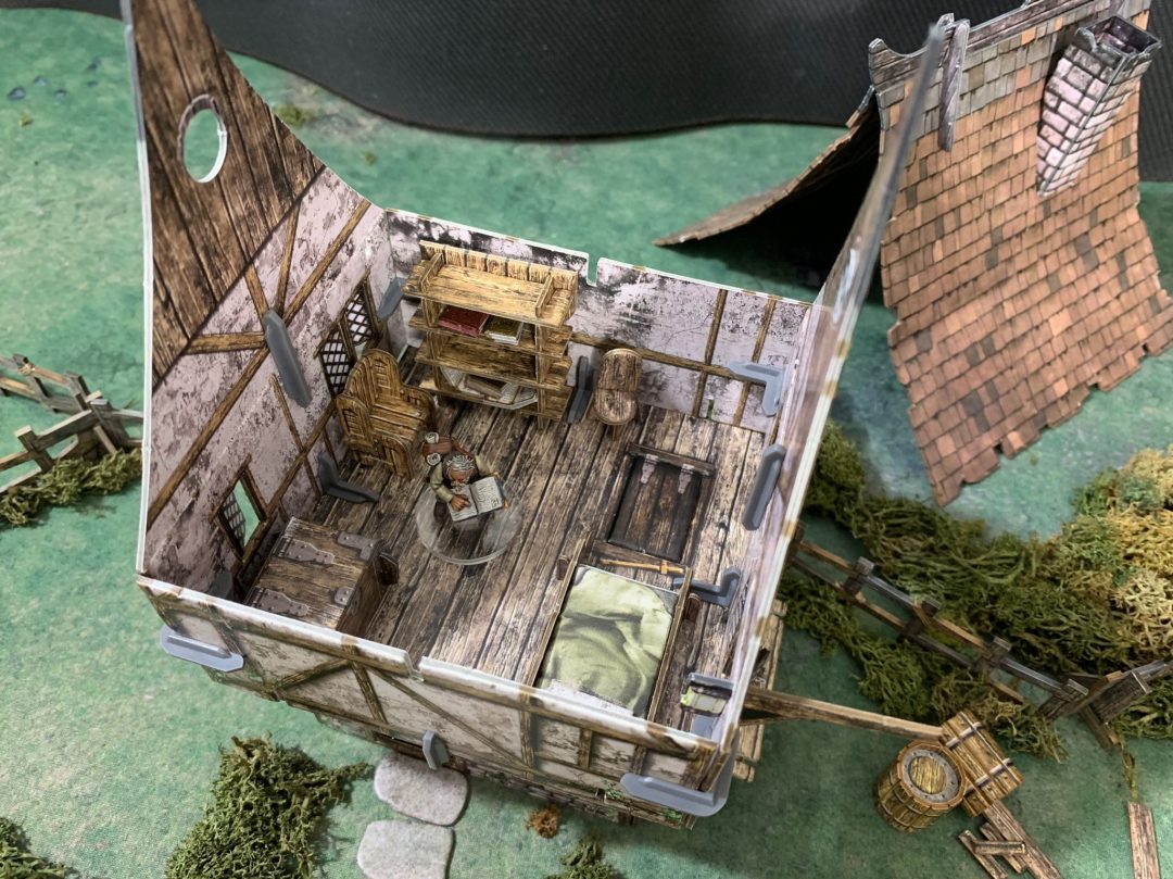 Detailed fantasy miniature house diorama. A robed figure reads at a table, with a bed and bookshelf inside.