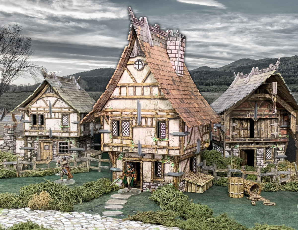 Tabletop diorama of a medieval village with three houses. A figure stands at the main door, another walks on green terrain.