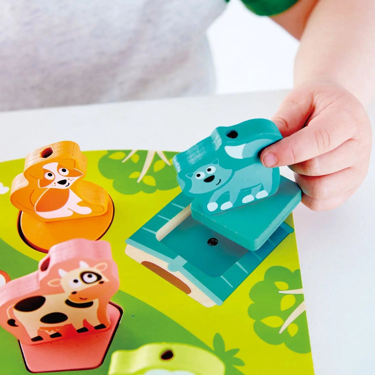 A child's hand places a teal animal puzzle piece onto a green wooden peg puzzle board.