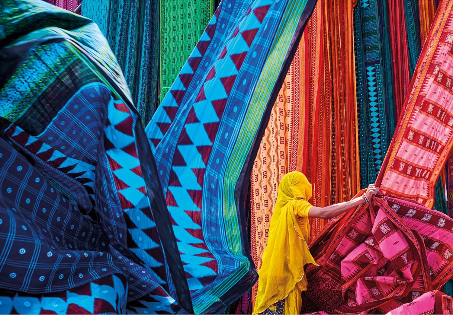 Person in yellow adjusts vibrant patterned textiles hanging like colorful drapes
