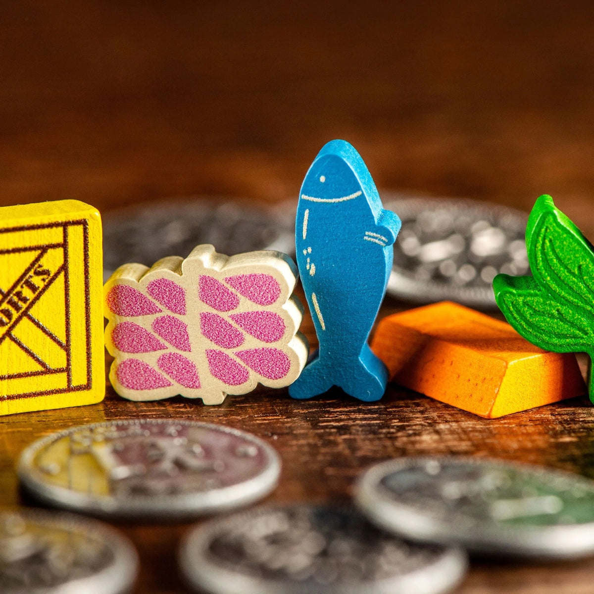 A close-up of colourful wooden Yokohama Premium Tokens including a yellow exports crate, a cream silk bundle, a blue fish, an orange copper bar, and a green tea leaf on a wooden table with blurred coins