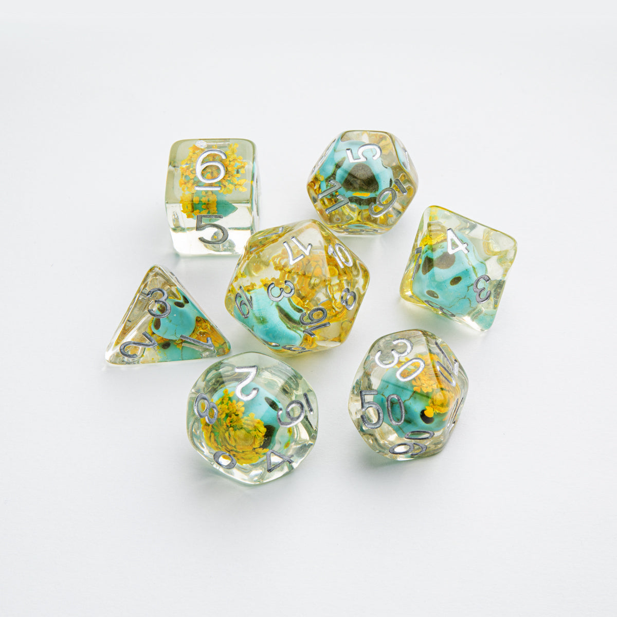 Seven clear resin polyhedral dice with embedded yellow flowers and blue-green stones, featuring silver numbers, on a white background