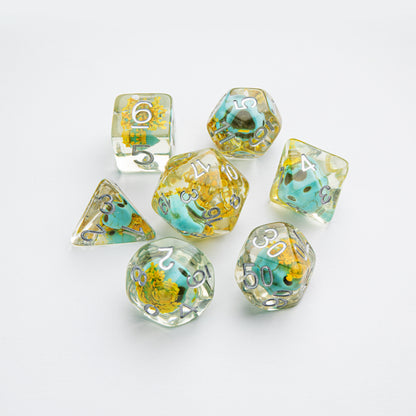 Seven clear resin polyhedral dice with embedded yellow flowers and blue-green stones, featuring silver numbers, on a white background