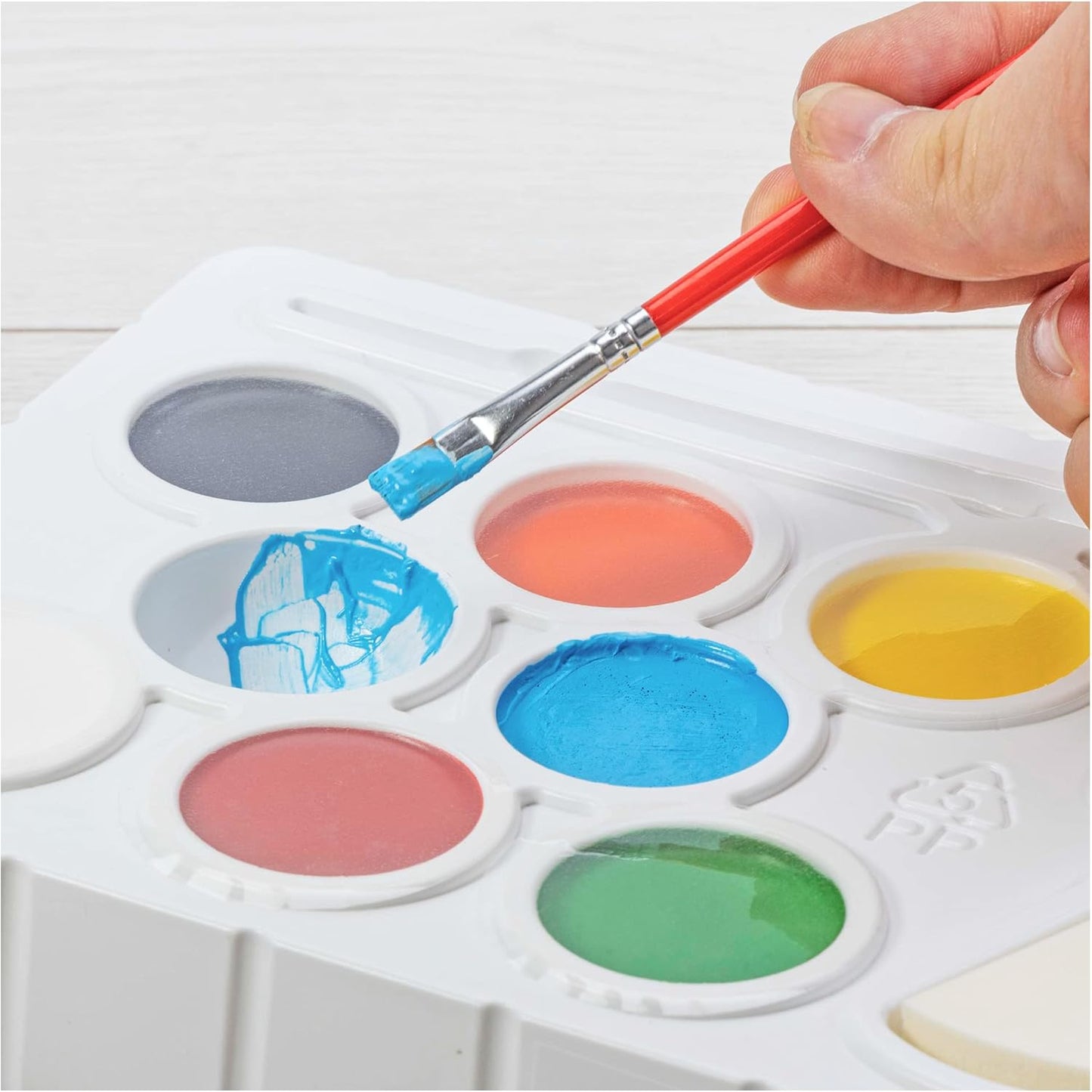 Hand dipping a red paintbrush into blue paint on a multi-colour art palette