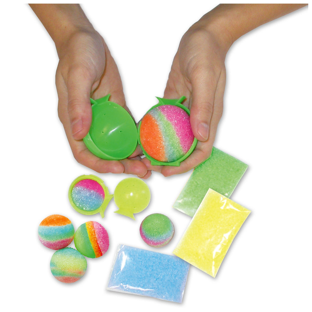 Hands hold an open green mold with a striped, glittery bath bomb, other colorful bath bombs and ingredient packets are displayed