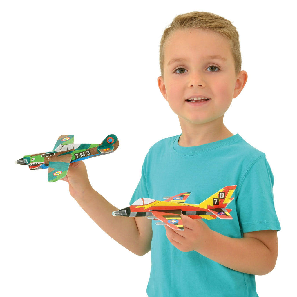 A smiling boy in a blue shirt holds a green propeller toy plane and a red-yellow toy jet