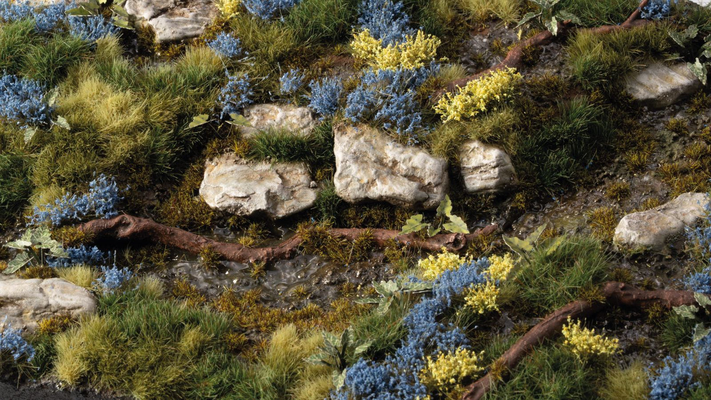 Realistic miniature landscape: rocky stream bed, diverse green grass, blue and yellow bushes, and a fallen branch
