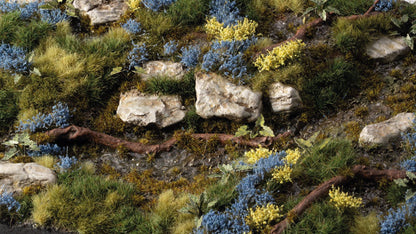 Realistic miniature landscape: rocky stream bed, diverse green grass, blue and yellow bushes, and a fallen branch