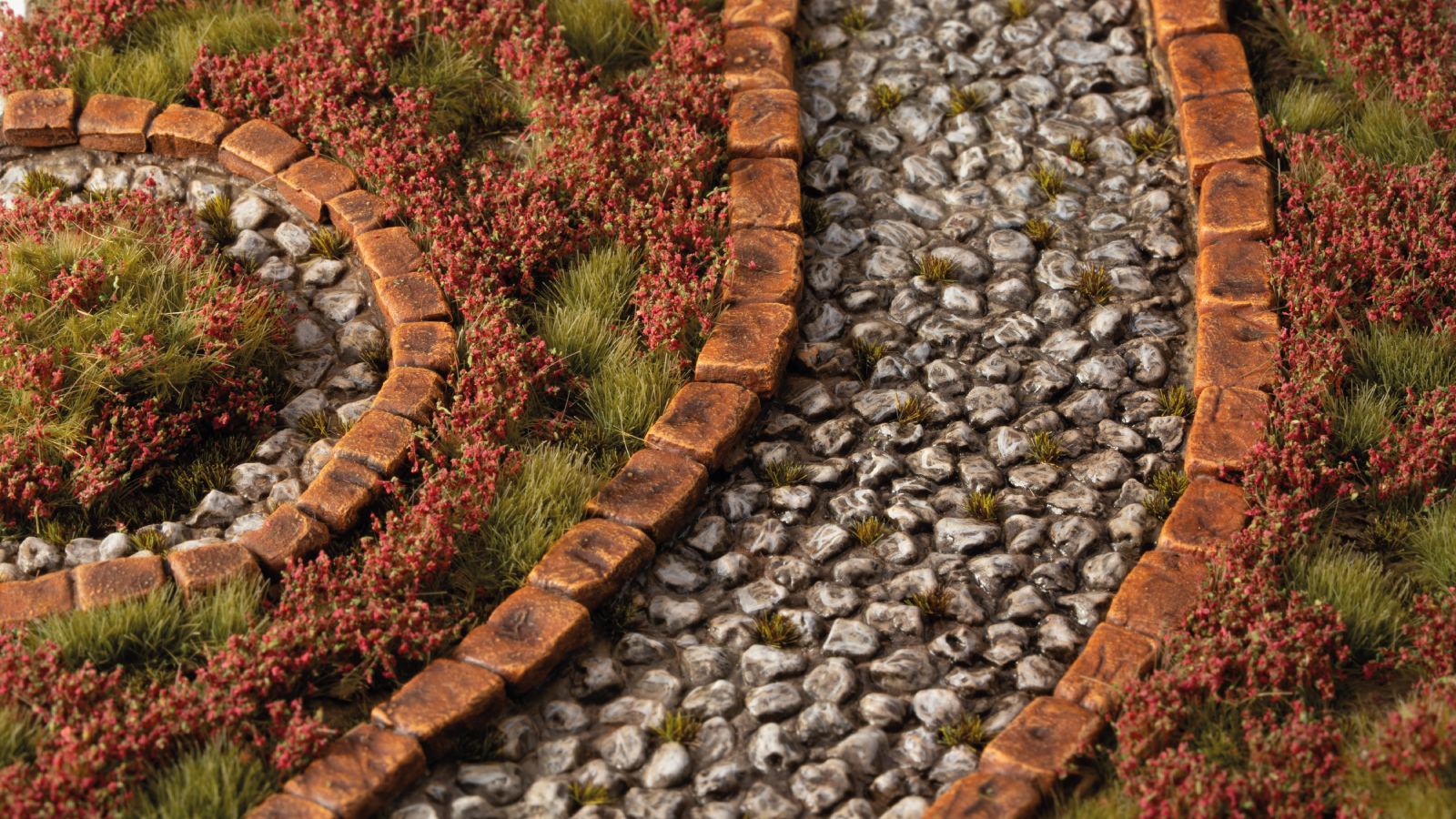 Miniature landscape model with two winding, brick-edged rocky beds, surrounded by green grass and small red plants
