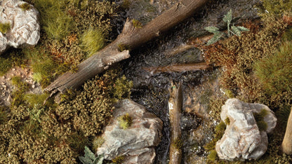Close-up of a miniature stream with flowing water, fallen logs, rocks, and patches of green moss and grass