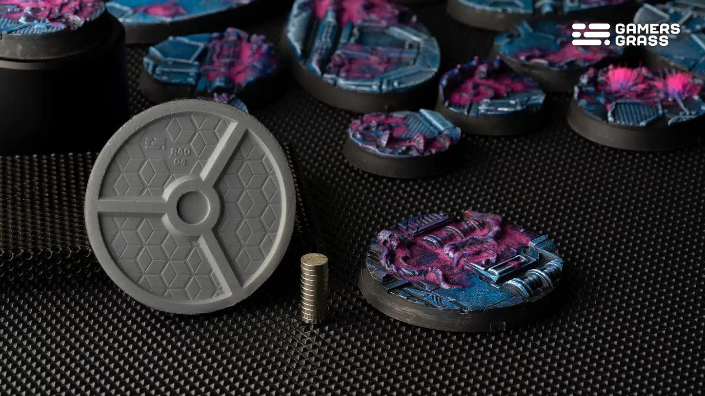 Underside of a grey miniature base with a magnet slot, a stack of small magnets and painted sci-fi terrain bases in blue and pink