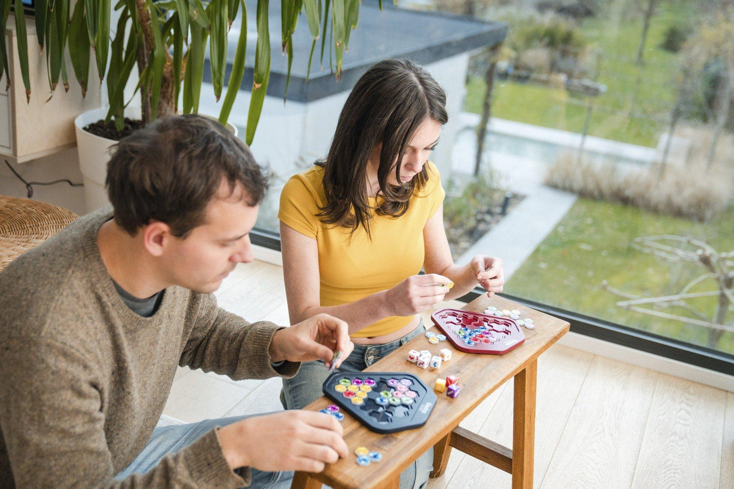 Two people intently play a board game with colorful pieces on a table by a window