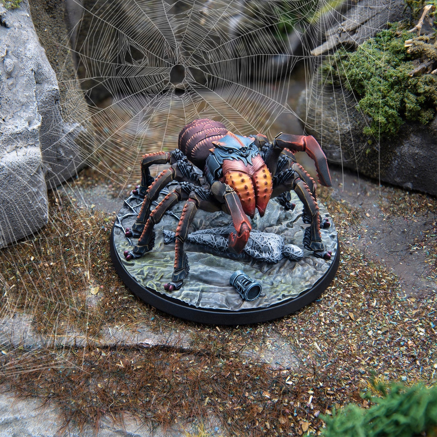 A detailed spider miniature with red-orange fangs, blue-grey head, and dark body, posed on a rocky base with a large spiderweb behind