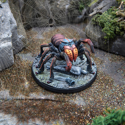 A detailed spider miniature with red-orange fangs, blue-grey head, and dark body, posed on a rocky base with a large spiderweb behind