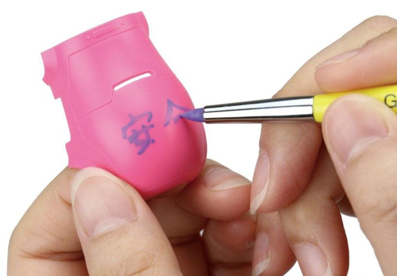 A hand uses a brush pen to write purple script on a pink plastic model piece