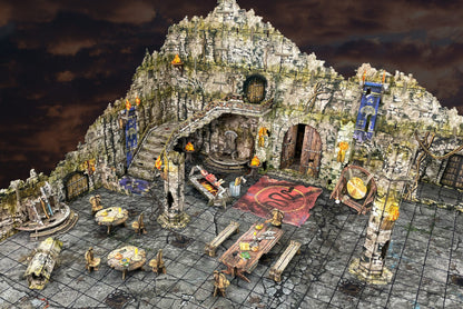 An intricate multi-level stone fortress tabletop terrain, featuring stairs, doors, tables and lit torches under dark clouds