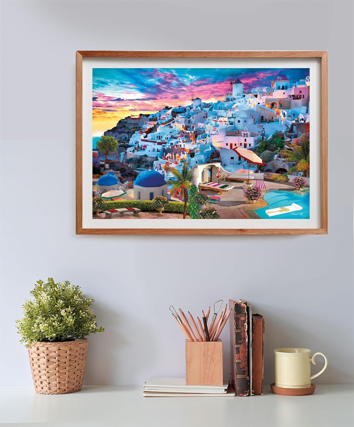 A vibrant framed print of a picturesque Greek village with white buildings, blue domes, and a windmill at sunset, above a desk.