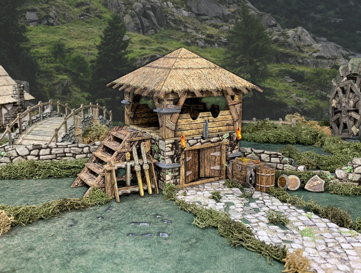 Detailed miniature diorama of a medieval thatched-roof workshop with stone base, lit torches, a cobbled path, bridge, and water wheel.