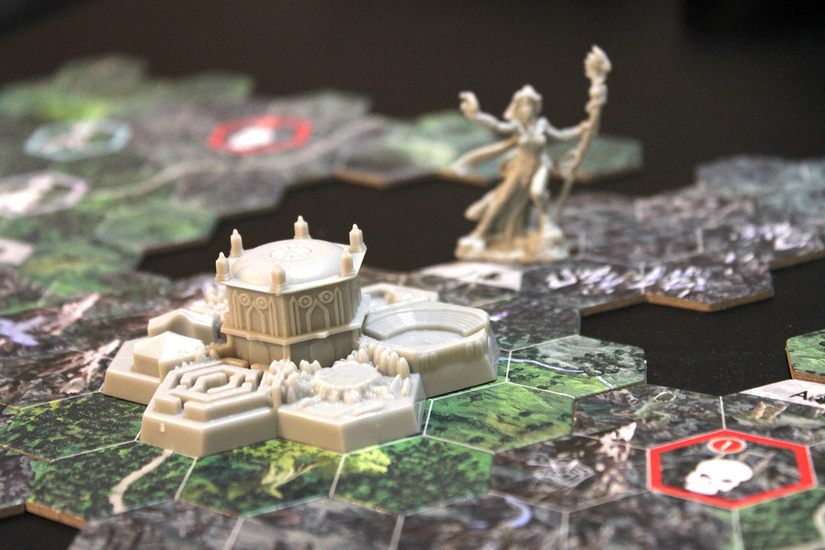 A close-up of a fantasy board game with hexagonal terrain tiles, a detailed beige temple-like building, and a blurry wizard miniature