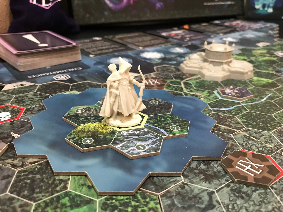 Close-up of a fantasy board game: a robed archer miniature stands on blue water hexes, surrounded by forest tiles