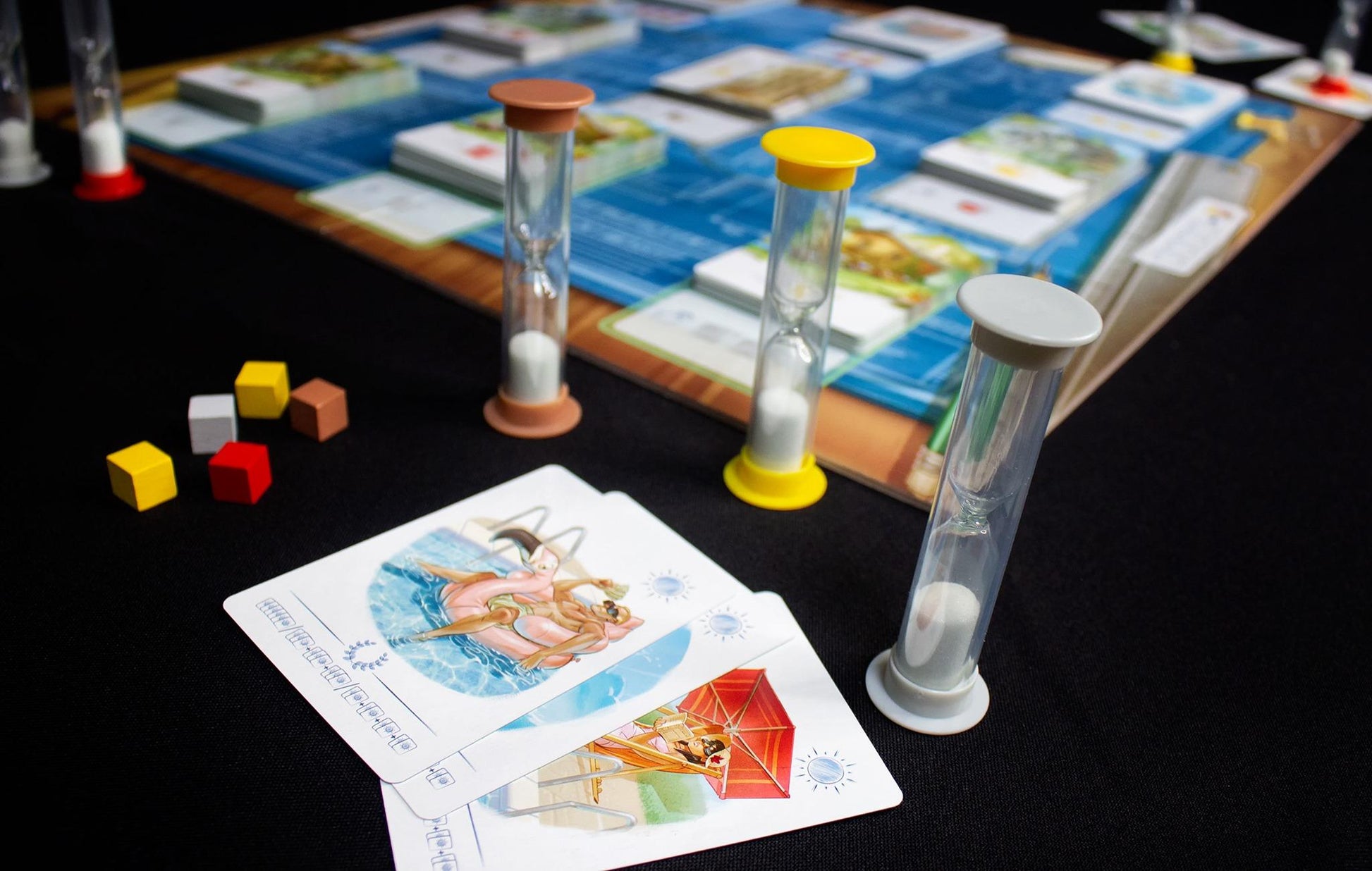 Board game pieces including three hourglass timers and five colored cubes next to two illustrated playing cards showing swimmers and a woman relaxing under a red umbrella