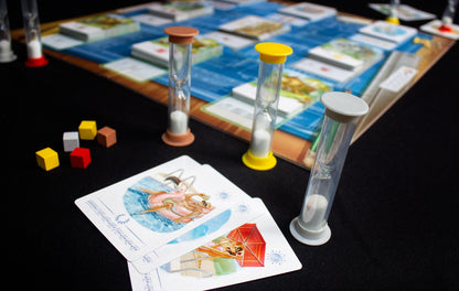 Board game pieces including three hourglass timers and five colored cubes next to two illustrated playing cards showing swimmers and a woman relaxing under a red umbrella