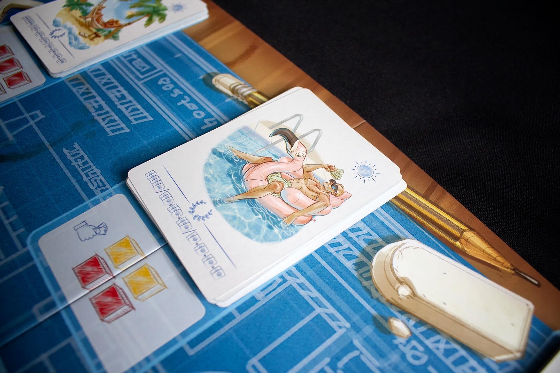 Board game cards featuring a person on a flamingo float two resource cards with red and yellow cubes and a pencil and eraser