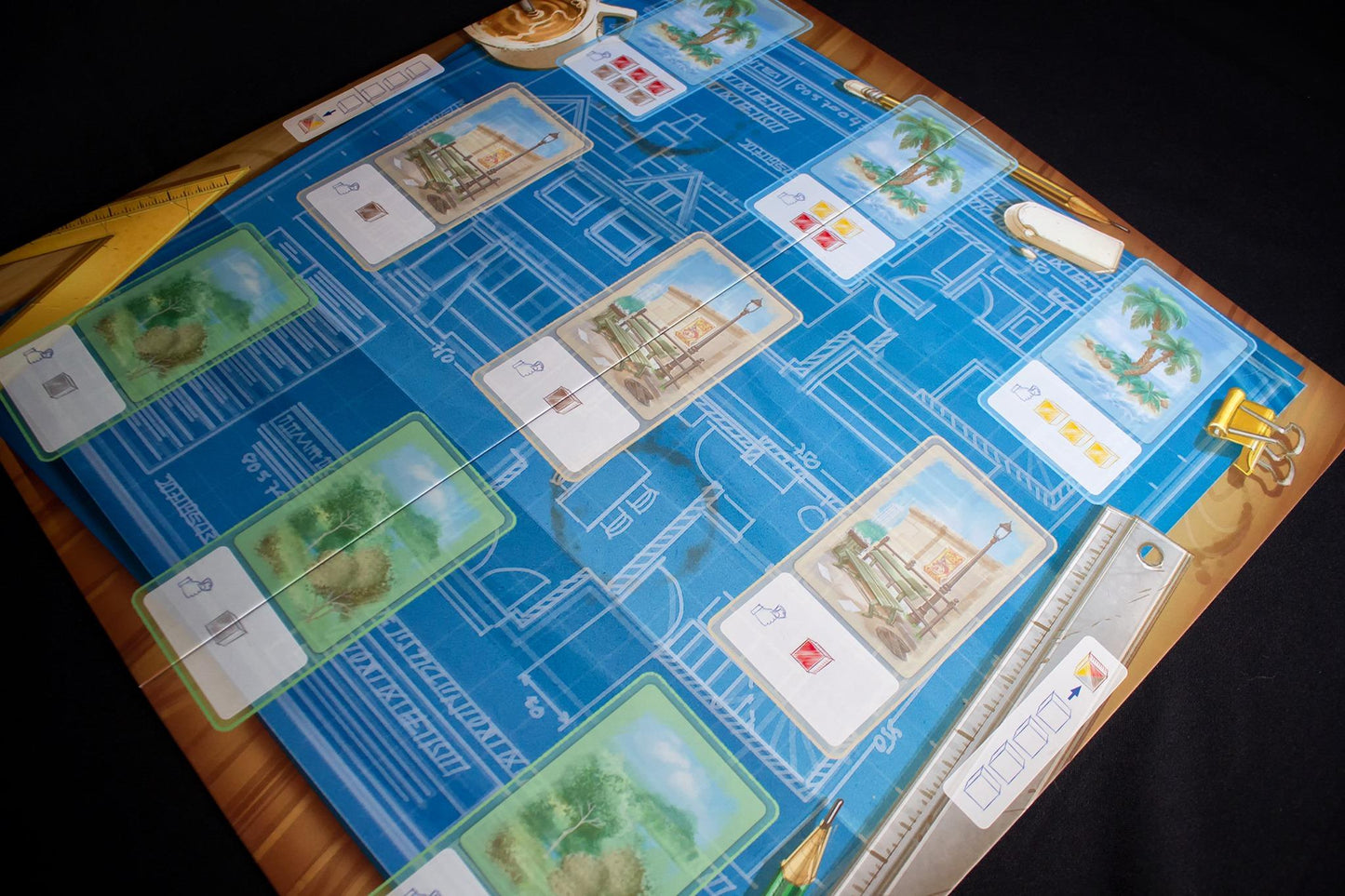 Blueprint style board game with cards featuring building projects landscaping and tropical island elements includes game pieces like rulers and pencils