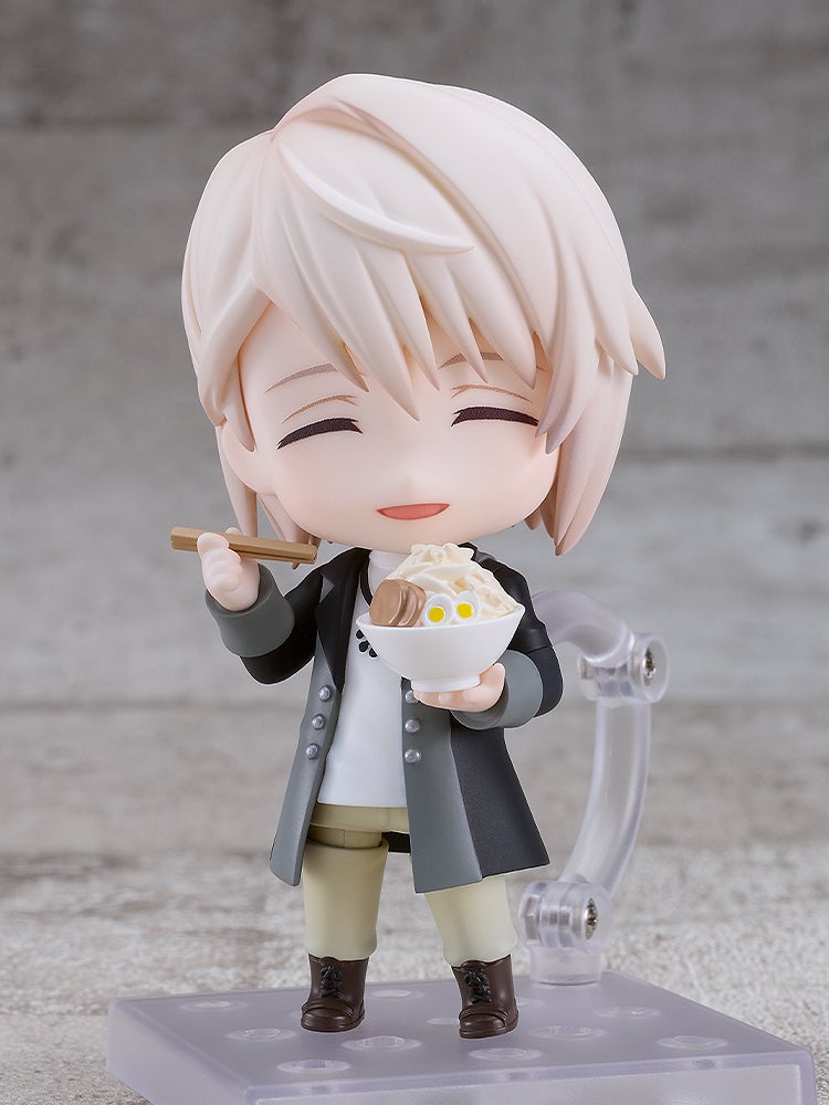 A cheerful Nendoroid figure with blonde hair smiles with closed eyes, eating ramen from a white bowl with chopsticks.