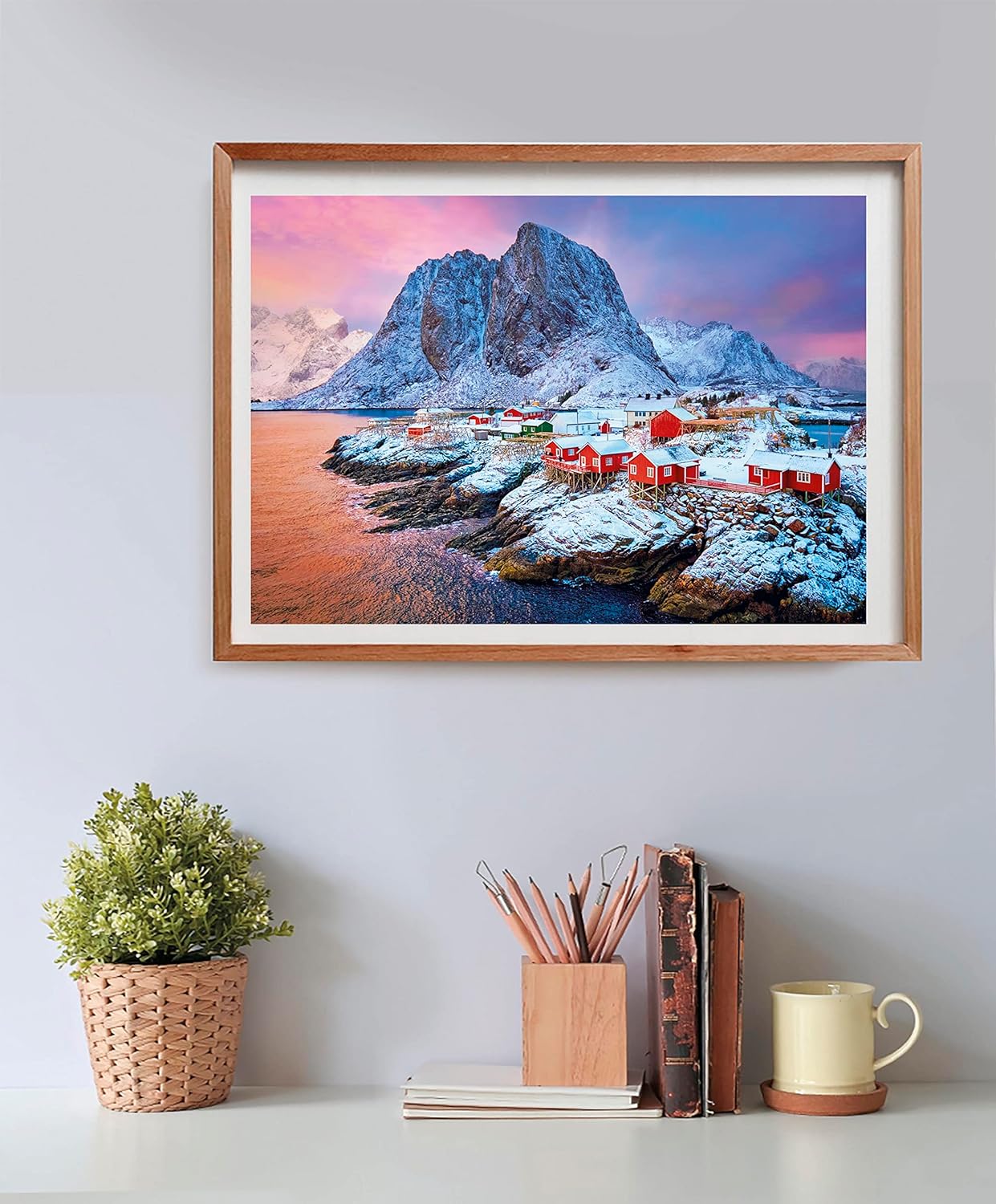 A framed print shows red houses on a snowy, rocky coast beside a fjord, backed by massive snow-covered mountains under a colorful sky.