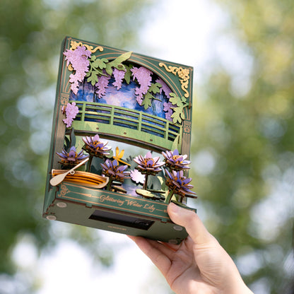 Layered wooden diorama of a water lily pond with a bridge, boat, and purple wisteria, held by a hand