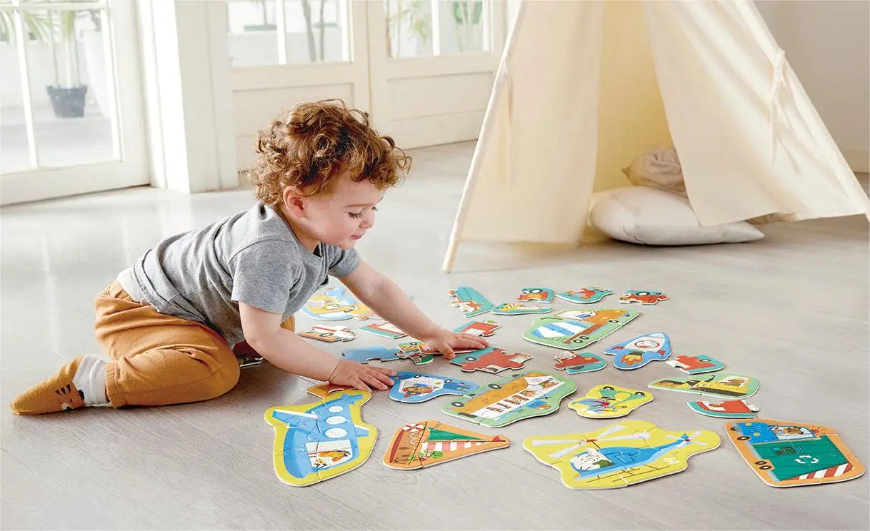 Toddler with curly hair kneels, assembling colourful vehicle puzzles on the floor, a play teepee behind.