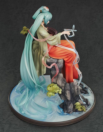 Hatsune Miku figure with long flowing teal hair that becomes water, seated on rocks wearing a green top and orange pants, holding an instrument and translucent ribbons