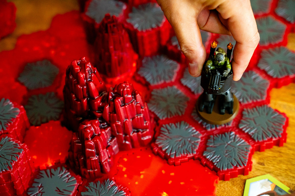 Hand places a green miniature figure on a board game map with red lava, jagged grey tiles, and rocky terrain