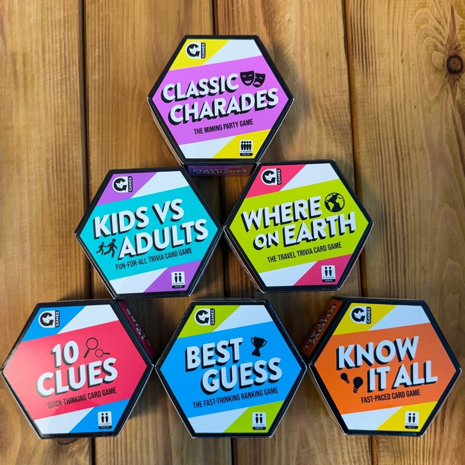 A collection of six colorful, hexagon-shaped party card game boxes stacked on a wooden background