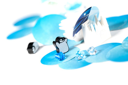 A wooden penguin game piece stands on blue ice floes, surrounded by clear crystal tokens and paper ice structures