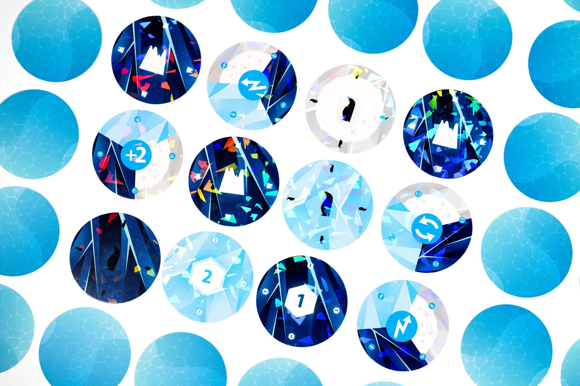 Circular blue-toned game tokens on white: some plain, some iridescent with numbers, arrows, mountains, and penguins