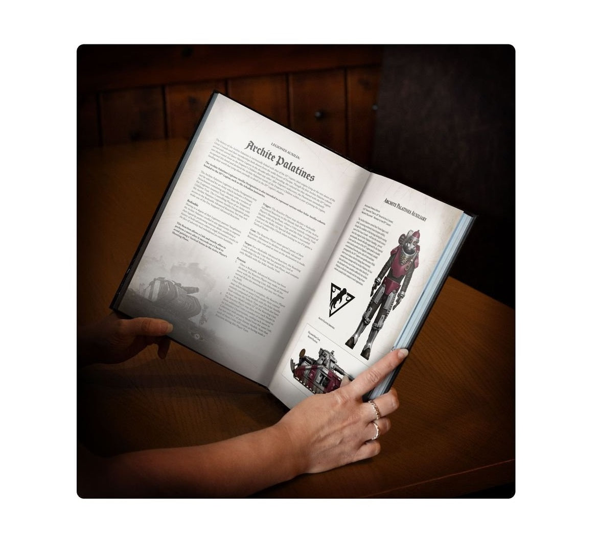 Hands hold an open rulebook titled "Archite Palatines," showing game text, a power-armored soldier, and a tank illustration