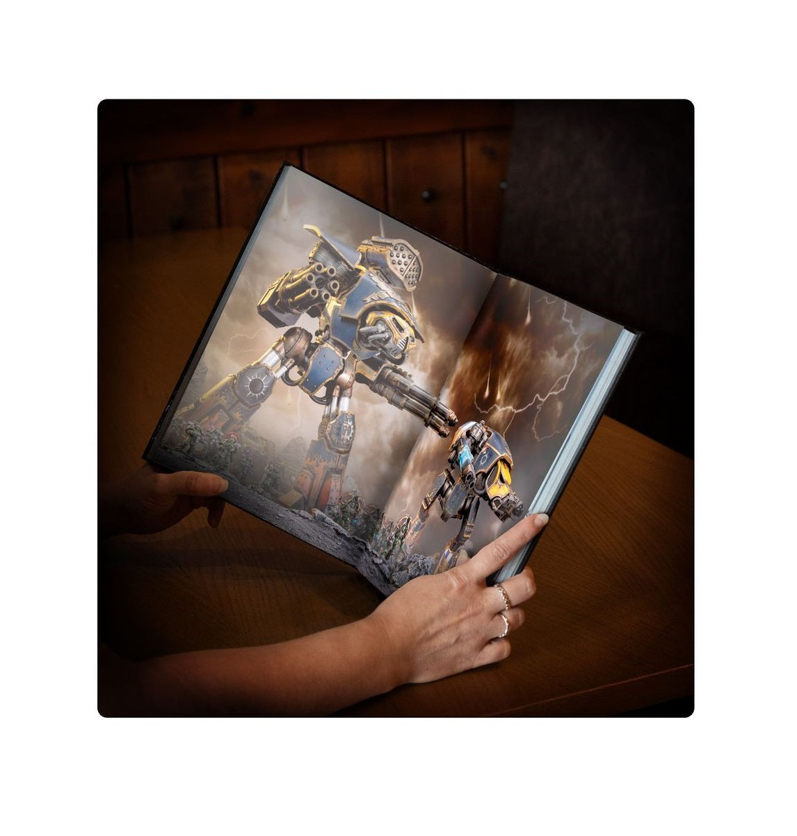 A person's hands hold an open book with an illustration of two giant mechs fighting amidst lightning