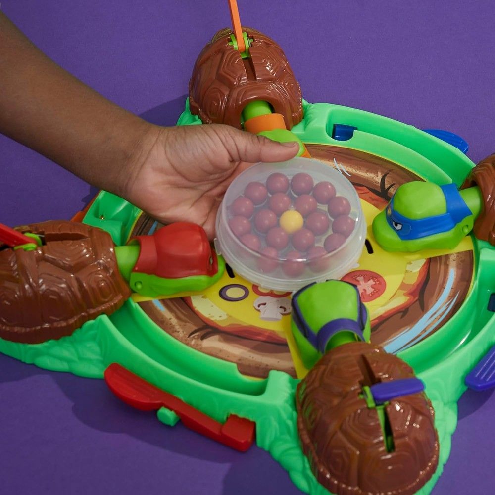 A hand holds a clear container of red and yellow balls over a Teenage Mutant Ninja Turtles board game with a pizza design