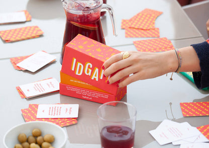 Hand holding a red "IDGA" game box on a table with scattered cards, a pitcher of red drink, and olives