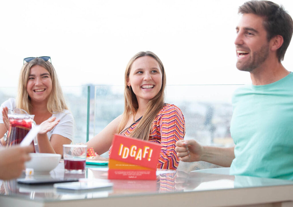 Smiling friends gather on a rooftop patio with drinks and a game box labeled 'IDGAF!' on the table