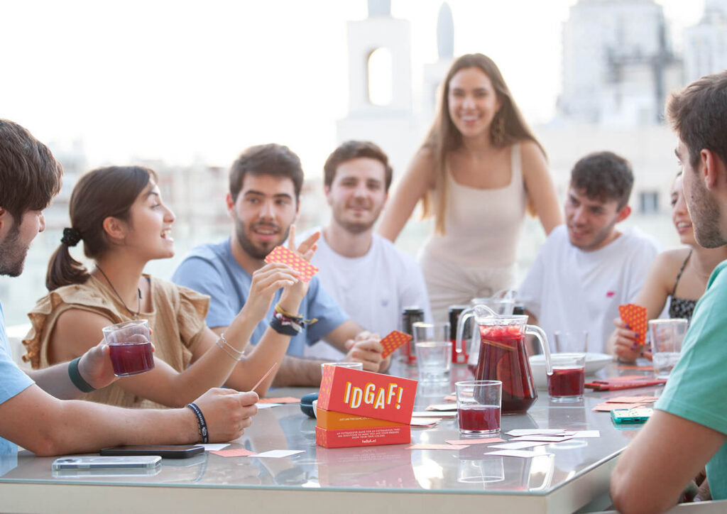 A group of friends play the "IDGAF!" card game with drinks on a sunny rooftop terrace