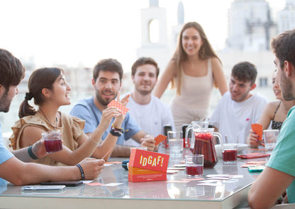 A group of friends play the "IDGAF!" card game with drinks on a sunny rooftop terrace