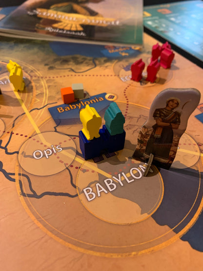 A close-up of a colorful board game with miniature yellow and teal game pieces on blue blocks a yellow and two gray and orange cubes near a card labeled Babylonia and a cardboard character piece