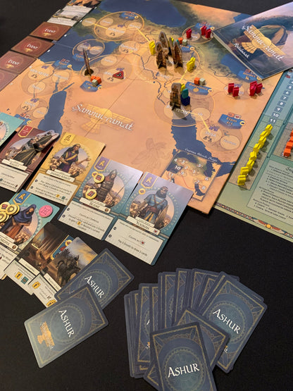 Overhead view of the Sammu-ramat board game with components including cards wooden pieces and the rulebook on a black surface