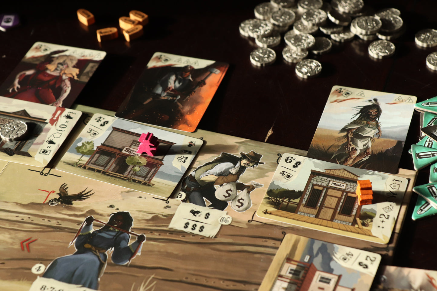 Close-up of a Western-themed board game, cards show characters, buildings, with pink and orange meeples, and silver coins